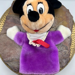 May include: A plush Minnie Mouse hand puppet. The puppet has a black head with large white eyes, a black nose, and a red mouth. It wears a purple bow and a purple dress with white sleeves. A white collar with a red ribbon completes the look.