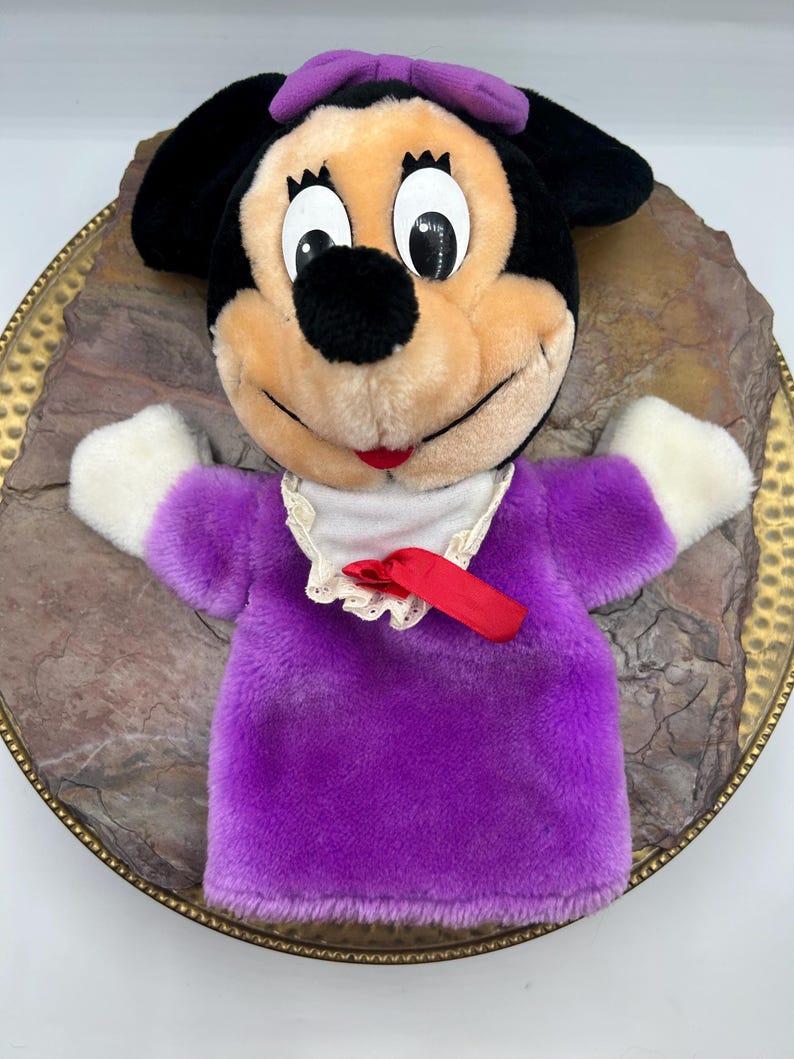 May include: A plush Minnie Mouse hand puppet. The puppet has a black head with large white eyes, a black nose, and a red mouth. It wears a purple bow and a purple dress with white sleeves. A white collar with a red ribbon completes the look.