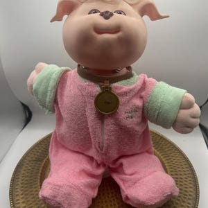 May include: A plush doll with a soft vinyl face, light pink skin, and tufted blonde hair. It wears a pink and light green terry cloth onesie with the text "MISS ME" embroidered on the chest. A brown collar features a round gold-colored tag.