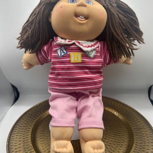 Könnte beinhalten: Eine Puppe mit braunen Wollhaaren, blauen Augen und einem sichtbaren Zahn. Die Puppe trägt ein rosa-weiß gestreiftes Oberteil mit weißem Kragen und drei kleinen quadratischen Aufnähern mit den Buchstaben A, B und C sowie eine rosa Hose.