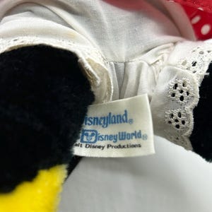 May include: Close-up of a plush toy featuring a red polka dot bow and a white dress with lace trim. The toy has black fur and a yellow beak. A tag reads "Disneyland" and "Disney World."