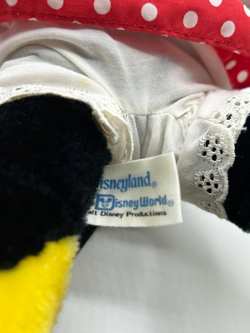 May include: Close-up of a plush toy featuring a red polka dot bow and a white dress with lace trim. The toy has black fur and a yellow beak. A tag reads "Disneyland" and "Disney World."