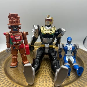 K&ouml;nnte beinhalten: Drei Actionfiguren auf einem goldfarbenen Teller. Die Figuren umfassen einen braunen Roboter mit dem Text "SSS", einen schwarzen und goldenen Power Ranger und einen blau-wei&szlig;en Power Ranger. Die Figuren sitzen.