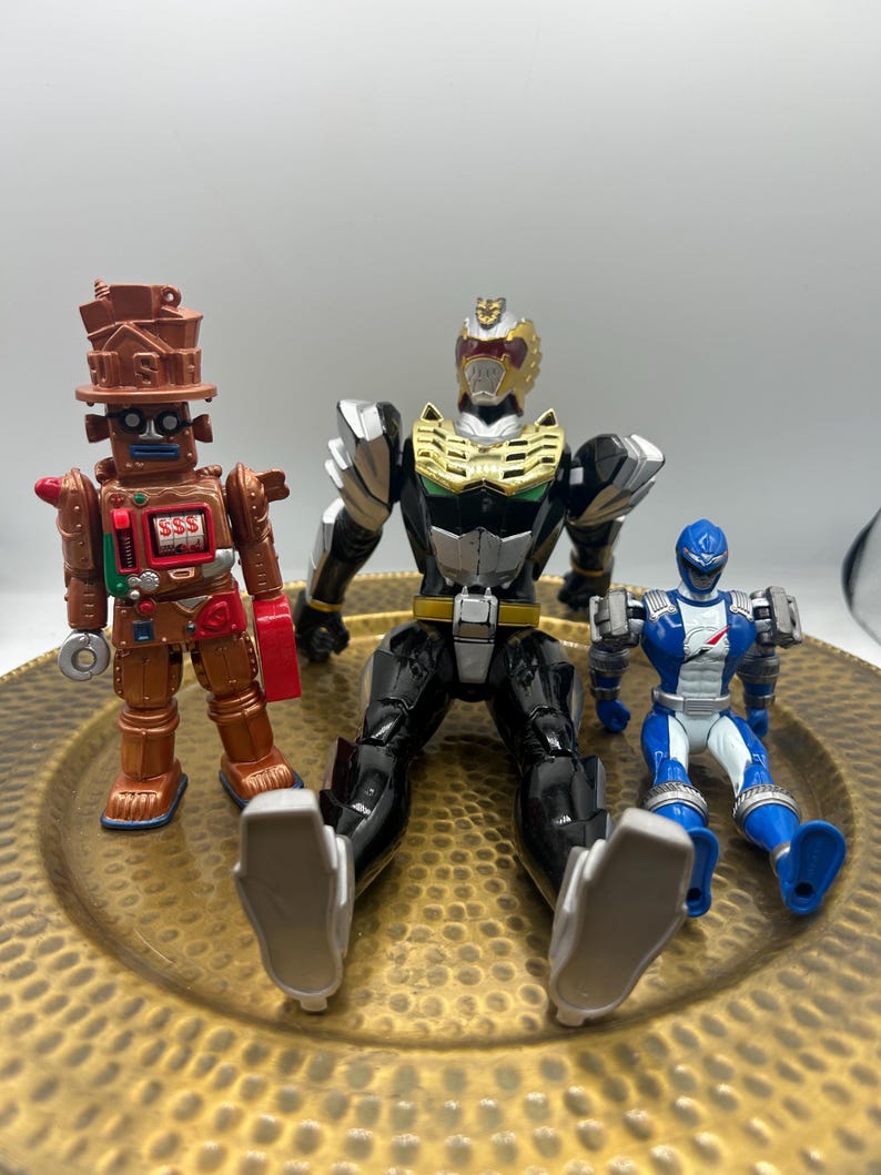 K&ouml;nnte beinhalten: Drei Actionfiguren auf einem goldfarbenen Teller. Die Figuren umfassen einen braunen Roboter mit dem Text "SSS", einen schwarzen und goldenen Power Ranger und einen blau-wei&szlig;en Power Ranger. Die Figuren sitzen.