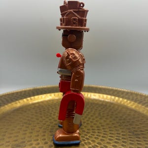 K&ouml;nnte beinhalten: Ein braunes und rotes Roboter-Spielzeug mit einem magnetf&ouml;rmigen Unterk&ouml;rper. Der Roboter hat einen Hut mit dem Wort "US" darauf, einen roten Knopf und einen silbernen G&uuml;rtel. Die F&uuml;&szlig;e des Roboters sind blau.