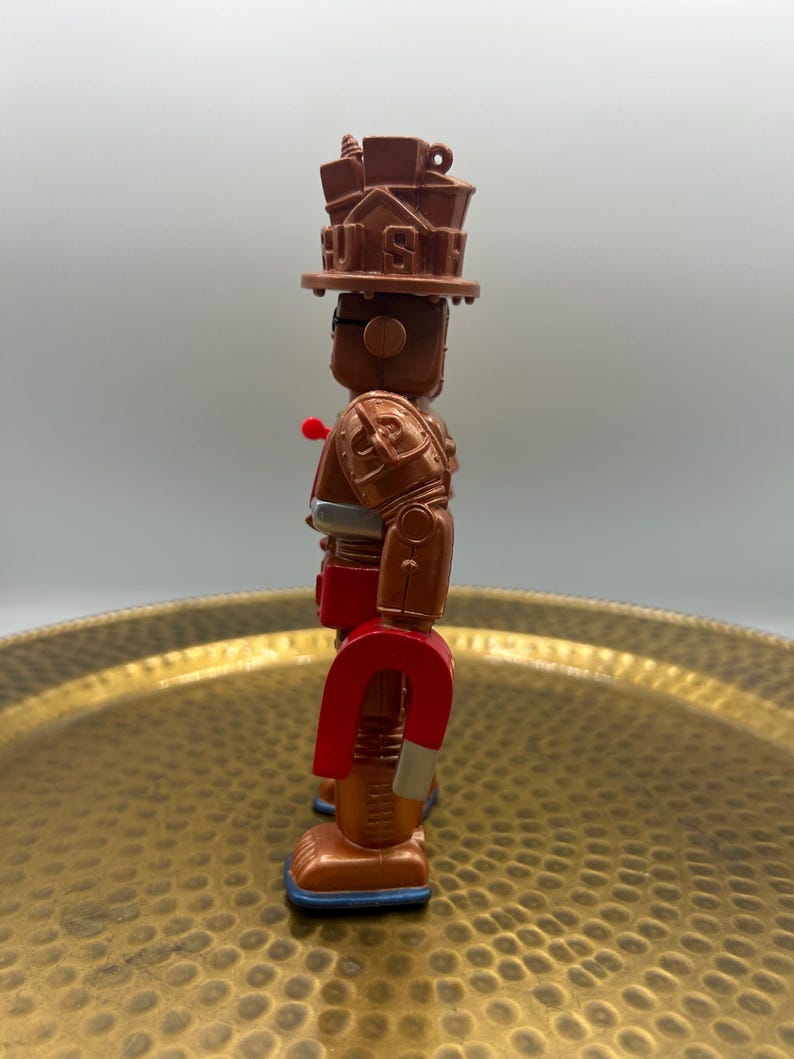 K&ouml;nnte beinhalten: Ein braunes und rotes Roboter-Spielzeug mit einem magnetf&ouml;rmigen Unterk&ouml;rper. Der Roboter hat einen Hut mit dem Wort "US" darauf, einen roten Knopf und einen silbernen G&uuml;rtel. Die F&uuml;&szlig;e des Roboters sind blau.