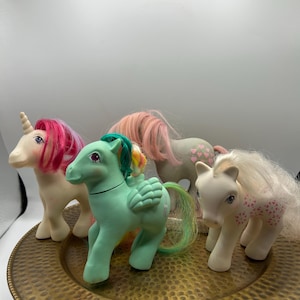 May include: Four plastic pony figurines with rooted manes and tails. One white unicorn with a pink mane, one mint green pegasus with a rainbow mane, one grey pony with pink heart symbols, and one white pony with pink heart patterns on its body.