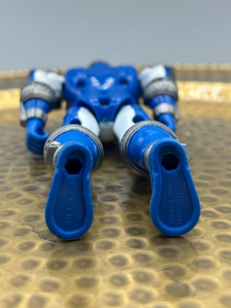 K&ouml;nnte beinhalten: Eine blaue und silberne Actionfigur mit beweglichen Gliedma&szlig;en. Die Figur hat ein V-f&ouml;rmiges Brustdesign und silberne Akzente an Armen und Beinen. Die F&uuml;&szlig;e haben eine einzigartige Form mit dem Text "BANDAI 2007 MADE IN CHINA".