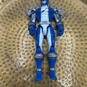 K&ouml;nnte beinhalten: Eine blau-wei&szlig;e Power Ranger-Actionfigur mit silbernen Akzenten an Schultern, Armen und Beinen. Die Figur hat einen Helm und einen wei&szlig;en Torso mit rotem Blitzdesign. Das Spielzeug liegt auf einer goldfarbenen Oberfl&auml;che.