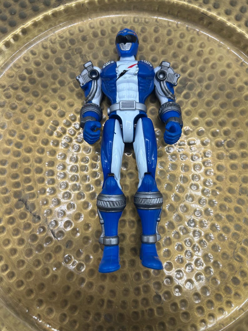 K&ouml;nnte beinhalten: Eine blau-wei&szlig;e Power Ranger-Actionfigur mit silbernen Akzenten an Schultern, Armen und Beinen. Die Figur hat einen Helm und einen wei&szlig;en Torso mit rotem Blitzdesign. Das Spielzeug liegt auf einer goldfarbenen Oberfl&auml;che.