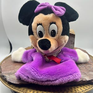 May include: A plush Minnie Mouse puppet with black ears, a tan face, and a purple bow. The puppet wears a purple dress with white gloves and a red ribbon. The puppet is sitting on a brown and gold decorative tray.