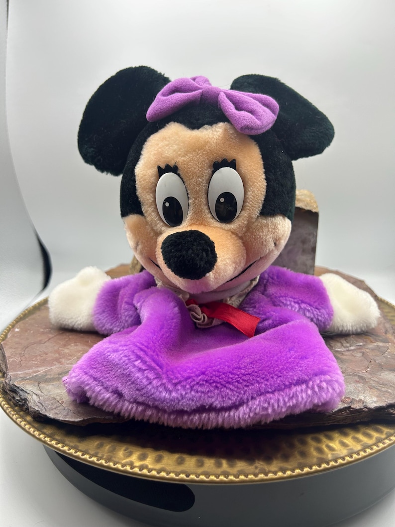 May include: A plush Minnie Mouse puppet with black ears, a tan face, and a purple bow. The puppet wears a purple dress with white gloves and a red ribbon. The puppet is sitting on a brown and gold decorative tray.