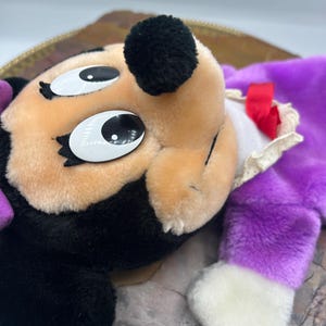 May include: A plush toy featuring a character with a black head, tan face, and large white eyes. The toy has a purple body, white gloves, and a red bow. The toy is lying on a decorative tray.