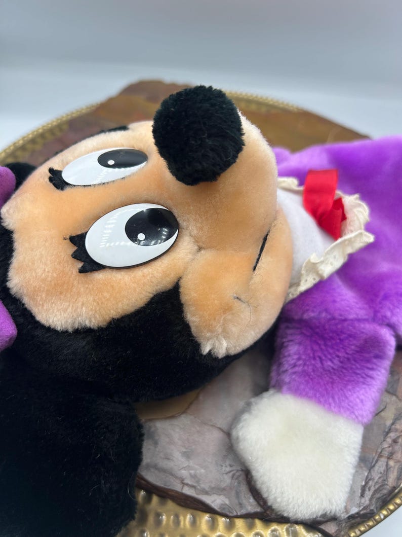 May include: A plush toy featuring a character with a black head, tan face, and large white eyes. The toy has a purple body, white gloves, and a red bow. The toy is lying on a decorative tray.