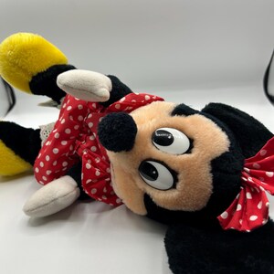 May include: A plush Minnie Mouse doll, featuring a black head and ears, a peach-colored face, and large, expressive eyes. She wears a red dress with white polka dots and a matching bow. Her yellow shoes and white gloves are also visible.