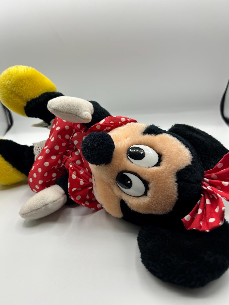 May include: A plush Minnie Mouse doll, featuring a black head and ears, a peach-colored face, and large, expressive eyes. She wears a red dress with white polka dots and a matching bow. Her yellow shoes and white gloves are also visible.