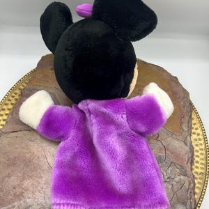 May include: A plush hand puppet featuring a black head with large ears and a purple bow. The puppet has a purple dress and white gloves. The puppet is resting on a decorative, gold-rimmed plate with a textured surface.