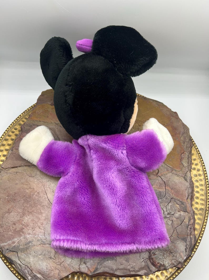 May include: A plush hand puppet featuring a black head with large ears and a purple bow. The puppet has a purple dress and white gloves. The puppet is resting on a decorative, gold-rimmed plate with a textured surface.