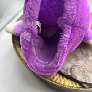 May include: A close-up of a purple plush toy with a white tip. The toy has a soft, fuzzy texture and a mesh interior. It is resting on a gold-colored tray.