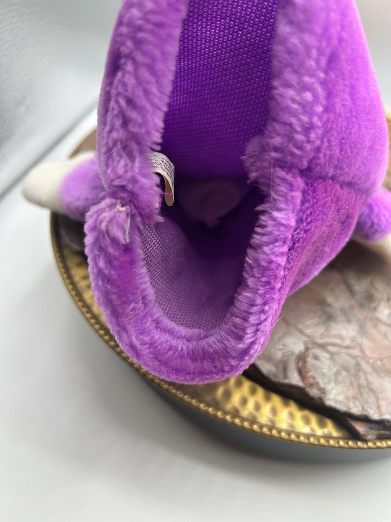 May include: A close-up of a purple plush toy with a white tip. The toy has a soft, fuzzy texture and a mesh interior. It is resting on a gold-colored tray.