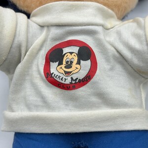Peut inclure: Une poup&eacute;e en peluche vintage de Mickey Mouse. La poup&eacute;e porte une chemise blanche avec un logo de cercle rouge qui dit "Mickey Mouse Club". La poup&eacute;e a un pantalon bleu et un visage bronz&eacute; avec des oreilles noires et un sourire rouge.