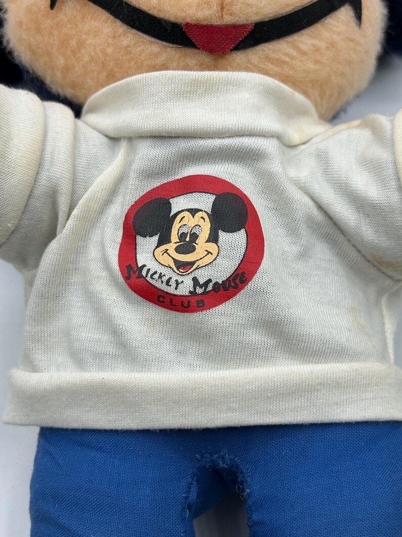 Peut inclure: Une poup&eacute;e en peluche vintage de Mickey Mouse. La poup&eacute;e porte une chemise blanche avec un logo de cercle rouge qui dit "Mickey Mouse Club". La poup&eacute;e a un pantalon bleu et un visage bronz&eacute; avec des oreilles noires et un sourire rouge.