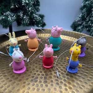 May include: A collection of seven small, colourful plastic character ornaments, each featuring a silver hanging string. The figures include various animal characters in shades of pink, yellow, blue, orange, and purple, arranged on a textured gold tray.