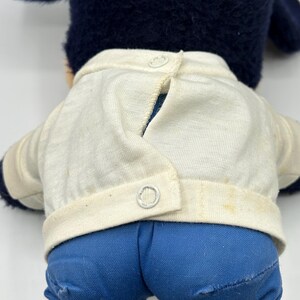 Peut inclure: Un jouet en peluche vintage avec une t&ecirc;te et des oreilles bleu fonc&eacute;, une veste blanche &agrave; deux boutons et un pantalon en jean bleu. Le dos du jouet est visible, montrant la fermeture boutonn&eacute;e de la veste et les d&eacute;tails du pantalon.