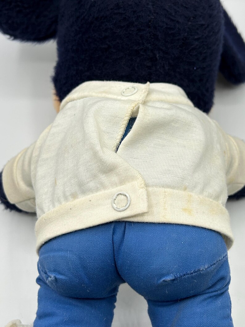 Peut inclure: Un jouet en peluche vintage avec une t&ecirc;te et des oreilles bleu fonc&eacute;, une veste blanche &agrave; deux boutons et un pantalon en jean bleu. Le dos du jouet est visible, montrant la fermeture boutonn&eacute;e de la veste et les d&eacute;tails du pantalon.