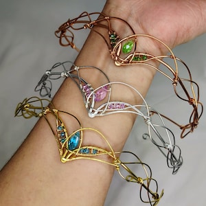 May include: Four wire-wrapped bracelets in copper, silver, and gold tones, each adorned with colorful gemstones. The bracelets are displayed on a person's arm, showcasing their intricate designs and craftsmanship.