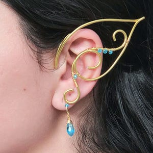 May include: Gold-toned ear cuff with a delicate, elven design. The cuff curves around the ear, adorned with small, light blue beads and a dangling teardrop-shaped blue gem. The jewelry is worn on the ear.