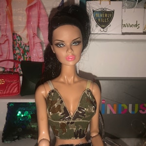 May include: A fashion doll with long black hair wearing a camouflage print crop top. The doll has a neutral expression and is wearing heavy eye makeup. The doll is sitting in front of a shelf with various items, including a red purse with a gold logo, a green purse, and a box with the word "INDUSTRY" printed on it.