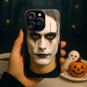 May include: Black phone case with a portrait of a person with white face paint, black eye makeup, and dark lipstick. The case has a black border and a camera module. The background includes a cup, a ghost, and a pumpkin.