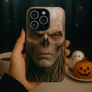 May include: A smartphone case with a detailed image of a pale, gaunt face with red eyes. The design has a spooky, Halloween theme. The case is held in a hand, with a blurred background.