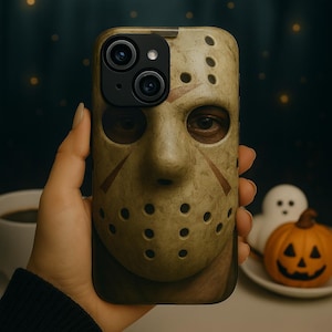 Puede incluir: Funda de teléfono con un diseño de máscara de hockey beige con detalles marrones y agujeros circulares. La funda está sujeta por una mano, con decoraciones de Halloween en el fondo. Los detalles de los ojos de la máscara son visibles.