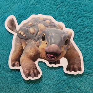May include: A close-up of a cartoon Ankylosaurus sticker. The dinosaur is brown and blue with a toothy grin and a large, round shell.