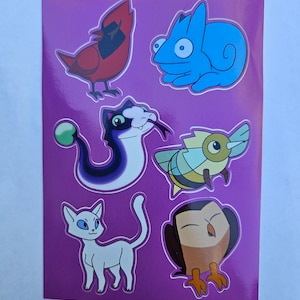 Puede incluir: Una hoja de pegatinas con ilustraciones de varios animales. Las pegatinas están sobre un fondo morado e incluyen un cardenal rojo, un camaleón azul, una serpiente blanca y negra, una abeja amarilla y marrón, un gato blanco y un búho marrón.