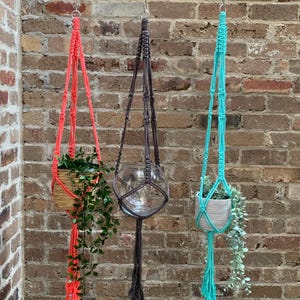 Op de afbeelding: Drie macramé plantenhangers, één in koraalroze, één in grijs en één in turquoise, hangen aan haken aan een bakstenen muur. Elke hanger heeft een plantenpot.