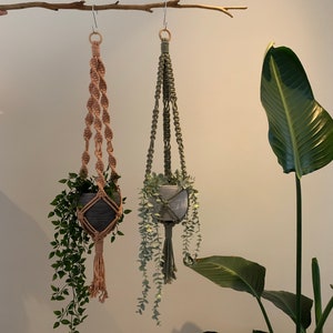 Op de afbeelding: Twee macramé plantenhangers met potten en planten die aan een tak hangen. De hanger aan de linkerkant is gemaakt van naturelkleurig macramé en de hanger aan de rechterkant is gemaakt van groen macramé.