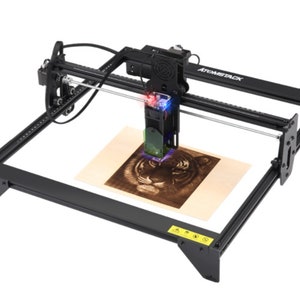 Puede incluir: Un grabador láser Atomstack negro está grabando activamente una imagen de tigre en un trozo de madera. La máquina tiene un marco rectangular con un cabezal láser móvil. La madera es de color marrón claro y el tigre grabado es detallado y marrón oscuro.