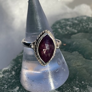 May include: A silver ring with a purple gemstone set in a marquise shape. The ring is on a white display stand.