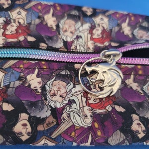 May include: A purple zippered pouch with a silver wolf head charm. The pouch is covered in a repeating pattern of characters from a fantasy novel.