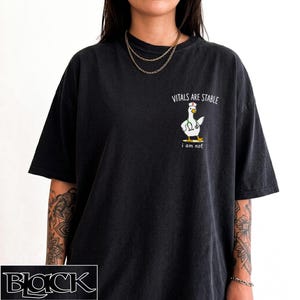 May include: A black short-sleeved t-shirt featuring a graphic of a white duck dressed as a nurse with a stethoscope. Above the duck, the text reads "VITALS ARE STABLE" and below the duck, the text reads "i am not" in white lettering.
