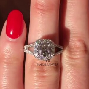 Puede incluir: Un anillo de compromiso de plata con un diamante central de forma cuadrada rodeado de diamantes más pequeños. El anillo tiene un diseño de caña dividida con dos garras a cada lado de la piedra central. El anillo está en un dedo con esmalte de uñas rojo.