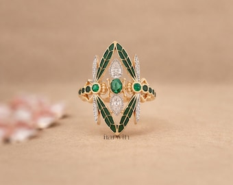 Op de afbeelding: Een gouden ring met een centraal smaragd- en diamantontwerp, geflankeerd door groene emaille vleugels. De ring heeft kleine smaragd- en diamantaccenten, waardoor een elegant en gedetailleerd sieraad ontstaat.