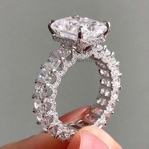 May include: A silver-toned ring featuring a large, rectangular, faceted clear stone set in a prong setting. The band is fully encrusted with smaller, square-cut clear stones arranged in a continuous eternity style, creating a highly reflective surface.