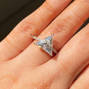 Könnte beinhalten: Ein silberner Ring mit einem großen, klaren, dreieckig geschliffenen Edelstein als Mittelpunkt. Das Band ist mit kleineren, klaren Steinen besetzt. Der Ring befindet sich an einem Finger und fängt das Licht ein.