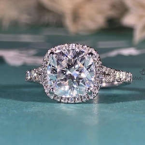 May include: A silver-toned ring featuring a large, cushion-cut centre stone surrounded by a halo of smaller, round-cut stones. The band is split and adorned with additional small, round-cut stones along the sides, creating a sparkling effect.