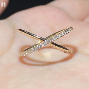 Op de afbeelding: Een goudkleurige crossover ring bestaande uit twee kruisende banden. De ene band is glad en gepolijst, terwijl de andere over de gehele lengte is ingelegd met een rij kleine, heldere, fonkelende steentjes. Het ontwerp vormt een X-vormig silhouet.