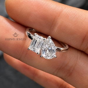 Peut inclure: Une bague argentée avec deux pierres transparentes proéminentes, l'une taille émeraude et l'autre taille poire, montées côte à côte sur un anneau poli. Le texte HARWIN JEWELS est imprimé à gauche près d'une petite icône florale.